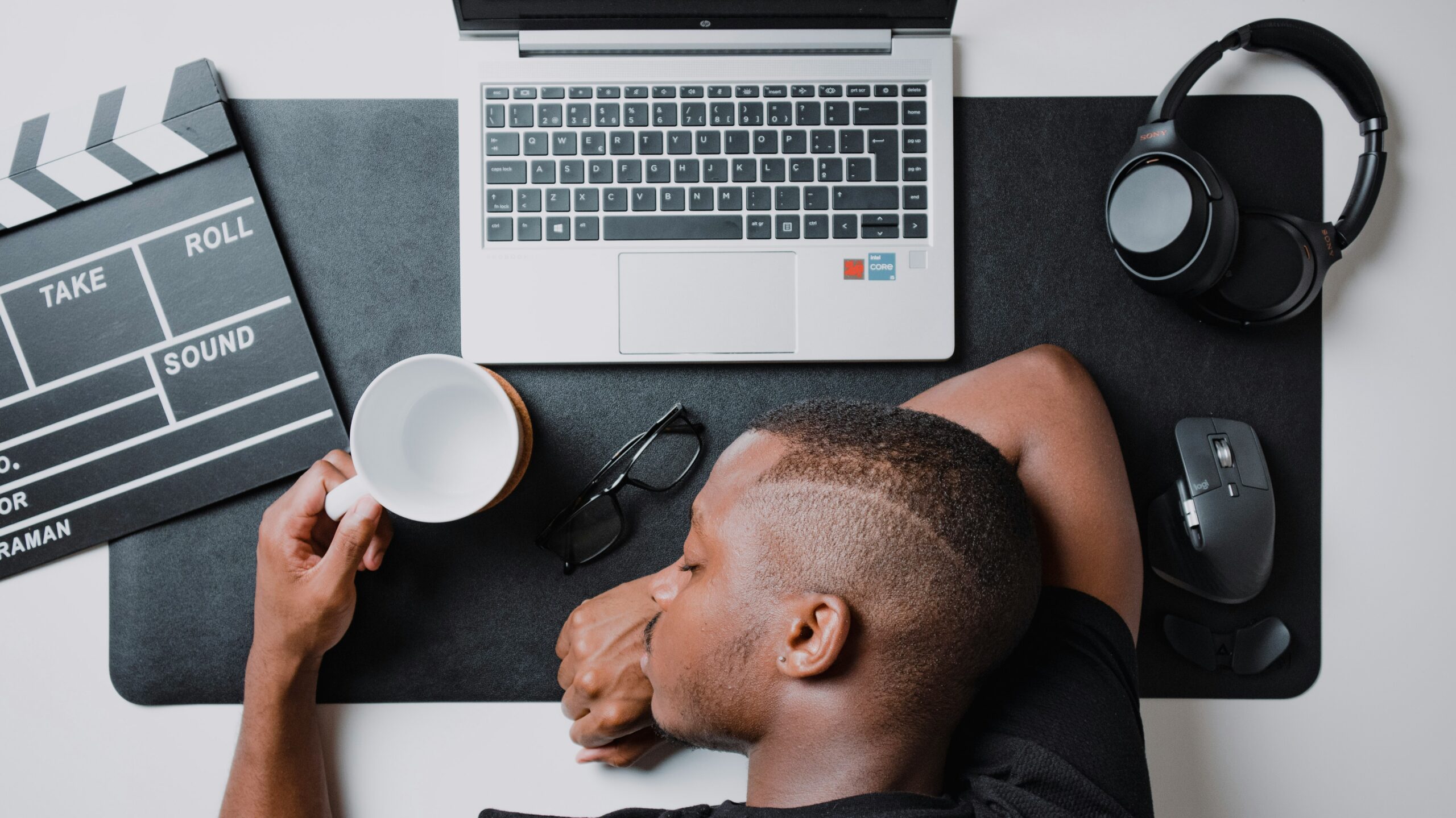 A man exhausted at his work desk, sleeping on a laptop with headphones, coffee mug, glasses, and clapperboard nearby, illustrating early signs of burnout at work.