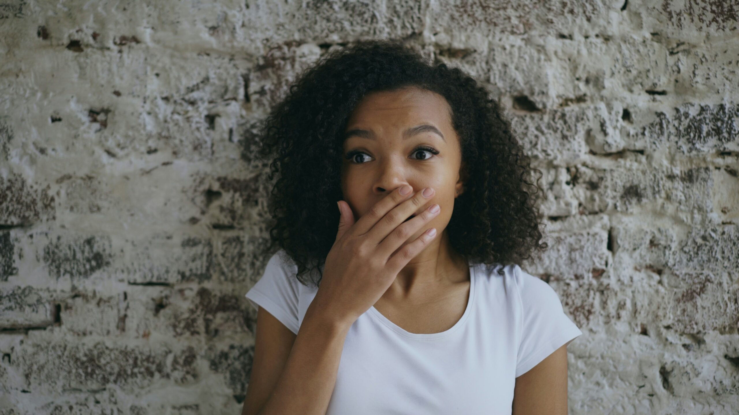 Woman standing against a brick wall with her hand near her mouth, suggesting a freeze response.